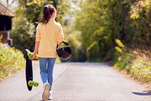 Rear View Of Girl With Skateboard Walking Along Country Road