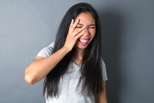 Young Beautiful Brunette Woman Wearing White T-shirt Over Grey Background Makes Face Palm And Smiles Broadly, Giggles Positively Hears Funny Joke Poses