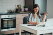 © Tijana - Smiling business woman sitting at kitchen desk and writing notes in a notebook while talking on a video call meeting with clients or coworkers on her laptop computer