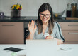 © Tijana - Smiling Businesswoman Waving And Say Hello To Colleagues At Web Conference Meeting On A Laptop Computer While Drinking Cup Of Coffee In The Kitchen