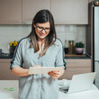 © Tijana - Happy business woman typing or reading business report online on her tablet while standing at the kitchen