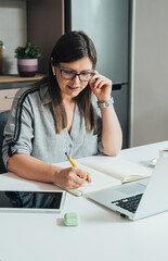 Smiling business woman sitting at kitchen desk and writing notes in a notebook while talking on a video call meeting with clients or coworkers on her laptop computer with wireless earphones