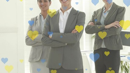 Wall Mural - Yellow and blue heart icons over three diverse businesswomen with arms crossed smiling at office