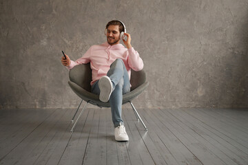 Wall Mural - Handsome man listening to music while sitting in comfortable armchair against gray wall.