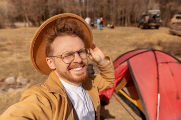 Wall Mural - Concept hiking travel adventure. Happy blogger man with hat making selfie photo portrait background red tent and mountains