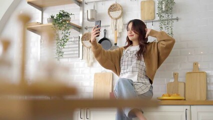 Poster - Pretty young asian female smile and sitting on counter kitchen room. She having fun taking selfie and video call talking with her friend