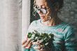 Close up portrait of young mature woman having care of a green plant at home in indoor leisure serene activity alone. Single lady enjoying life and apartment. One female people wearing eyewear indoor