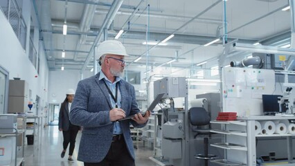 Poster - Portrait of a Bearded Middle Aged Chief Engineer Walking in a Factory Facility, Wearing Casual Suit and a White Hard Hat. Heavy Industry Specialist Working on Laptop Computer.