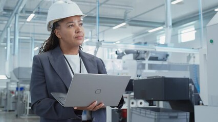 Fototapete - Portrait of a Black Female Engineer in Hard Hat Standing and Using Laptop Computer at Electronic Manufacturing Factory. Technician Thinking About Daily Tasks and Working on Project Pipeline.