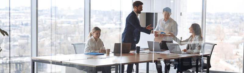Wall Mural - People standing near table, team of young businessmen working and communicating together in office.