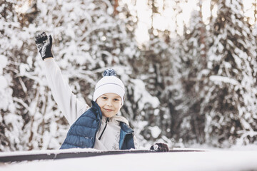 Wall Mural - Teenager boy looking out of car window traveling in winter snowy forest. Road trip adventure and local travel concept. Happy child enjoying car ride. Christmas winter holidays and New year vacation