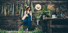 Attractive Girl Wearing Straw Hat And Blue Denim Dungarees Relaxing Near Wooden Old Summerhouse Wall On Sunny Day.