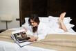 © Dragosh - Cheerful brunette young woman relaxing on a bed. Young beautiful woman wearing white bathrobe lying on bedroom.
