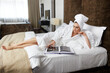 © Dragosh - Cheerful young woman in white bathrobes with towel on hier head, browse a magazines, lying down on bed.
