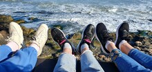 The Girls Are Resting On The Sea. Women's Legs In Sneakers On The Background Of The Sea. Vacation At Sea Is Not In Season. Female Friendship.
