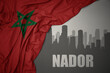 abstract silhouette of the city with text Nador near waving colorful national flag of morocco on a gray background.