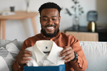 Wall Mural - African American Man Holding New Shirt Unpacking Shopper Bag Indoors