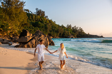 Young beautiful couple, in white clothes, walk along the sand along the coastline, hold hands, against the backdrop of the sunset.