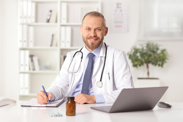 Canvas Print - Smiling young male doctor sitting in an office and looking at camera and writing