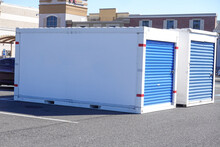 Two White Storage Pods With  Blue Doors On A Asphalt Road In Front Of Some Stores