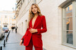 © Svetlana Sokolova - Beautiful smiling woman  in  elegant red velvet suit holding purse and  posin outdoor in old european city.    Blond wavy hairs, perfet skin, full lips.