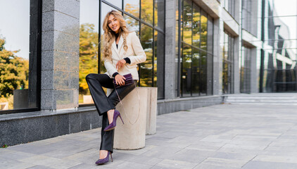 Wall Mural - Outdoor photo of sucsessful blond woman with perfect wavy hairs in casual  outfit with luxury purse posing over modern business center.