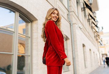 Wall Mural - Beautiful smiling woman  in  elegant red velvet suit holding purse and  posin outdoor in old european city.    Blond wavy hairs, perfet skin, full lips.