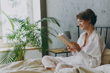 Side view young calm happy woman wear white shirt pajama she lying in bed read book study rest relax spend time in bedroom lounge home in own room hotel wake up dream be lost in reverie good mood day