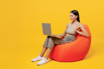 Full body young happy latin woman 30s she wearing basic beige tank shirt sit in bag chair hold use work on laptop pc computer waving hand talk on video call isolated on plain yellow backround studio