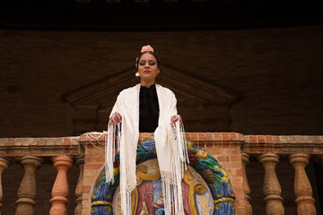 A beautiful teenage flamenco dancer with brown hair stands on a balcony wearing a shawl embroidered with fringes and performs flamenco dance postures. World Cultural Heritage