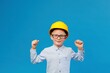 © nazariykarkhut - Cute boy construction worker in a yellow hard hat stands in an indoor studio on a blue background and shows satisfaction with the work done.