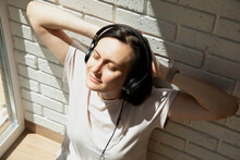 A Woman Near The Window Wearing Black Headphones And Listening To Music With Her Eyes Closed. View From Above, Close Up