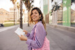 © Look! - Pretty young caucasian girl turns around looking away walking around European city. Brown-haired woman holds notebook, wears shirt and string bag. Relaxed lifestyle, concept