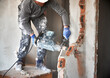 Close up of male builder in workwear drilling wall with hammer drill. Man worker using drill breaker while destroying wall in apartment under renovation. Demolition work and home renovation concept.
