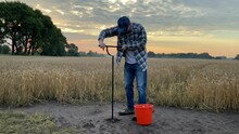 Male Agronomist Taking Sample With Soil Probe Sampler On Background Of Grain Field At Dawn. Farmer Using Drilling Tool For Soil Sampling At Morning Outdoors. Environmental Protection, Organic Research
