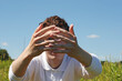 Young man hands and eye behind hands on a sky and grass background. Photo was taken 29 June 2022 year, MSK time in Russia.