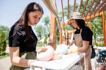 Wall Mural - Young brunette girl buyer with long hair and woman seller in apron and hat on either side of the counter with goods at the street fair in summer