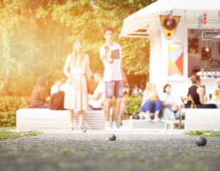 Wall Mural - Couple playing petanque guy through a ball above trees in city park outdoor activity sunshine