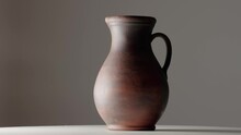 Brown Earthenware Jug With A Handle Spinning In A Dark Studio. An Old Jar From Clay Rotating On A White Table Isolated On Dark Gray Background. Handmade Ancient Pitcher For Drinks And Food.
