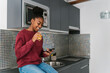 © Ezequiel Giménez/Stocksy - Cheerful black woman surfing smartphone in kitchen