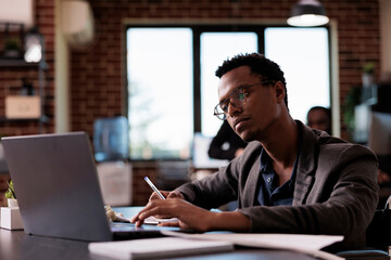 Wall Mural - African american man with impairment analyzing online report on laptop, planning project strategy in disability friendly office. Entrepreneur with health condition sitting in wheelchair at job.