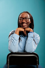 Wall Mural - Joyful pleased young adult person with confident smile and casual looking. Excited cheerful african american pretty attractive woman sitting on chair while smiling at camera on blue background.