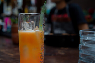 Wall Mural - Close up of a glass of cold juice with an ice cube and a green leaf decoration. Drink on a bar counter with the bartender in the background. Yellow drink in a night club.