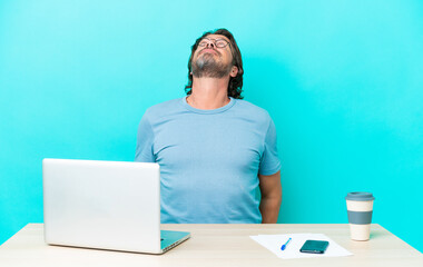 Senior dutch man in a table with a laptop isolated on blue background and looking up