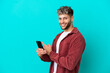 © luismolinero - Young handsome caucasian man isolated on blue background using mobile phone and pointing back