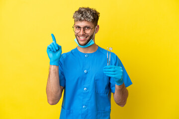 Wall Mural - Dentist caucasian man holding tools isolated on yellow background showing and lifting a finger in sign of the best