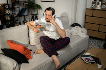 A young man thoughtfully reads text in book in English. He has headphones on head. He rested head on his arm. There is plaid and telephone on the sofa.