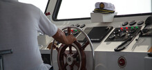 The Captain Of The Ship Holds The Helm With His Hand And The Captain's Cap Lies Nearby. White Cockpit With Brown Steering Wheel.
