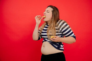 Overweight adipose woman with naked belly in striped clothes holding and eating potato chips. Unhealthy fattening food