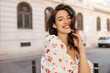 © Look! - Positive young caucasian girl smiles teeth looking at camera on background of city. Brunette with wavy hair wears white blouse in summer. Concept of enjoying weekend, vacation